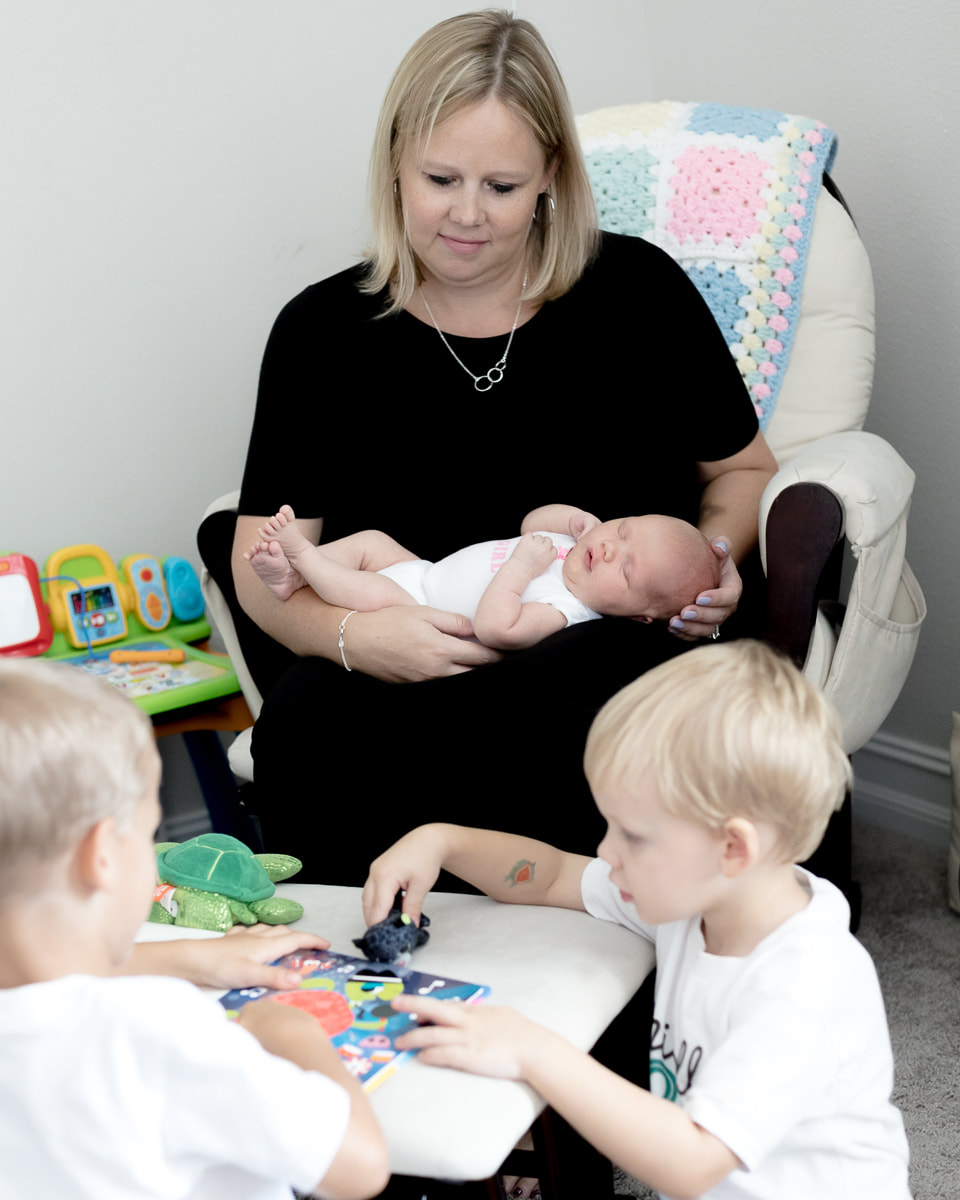 Mom in nursery holding newborn with older brothers looking at books, lifestyle newborn photography, Lakewood Ranch, Florida