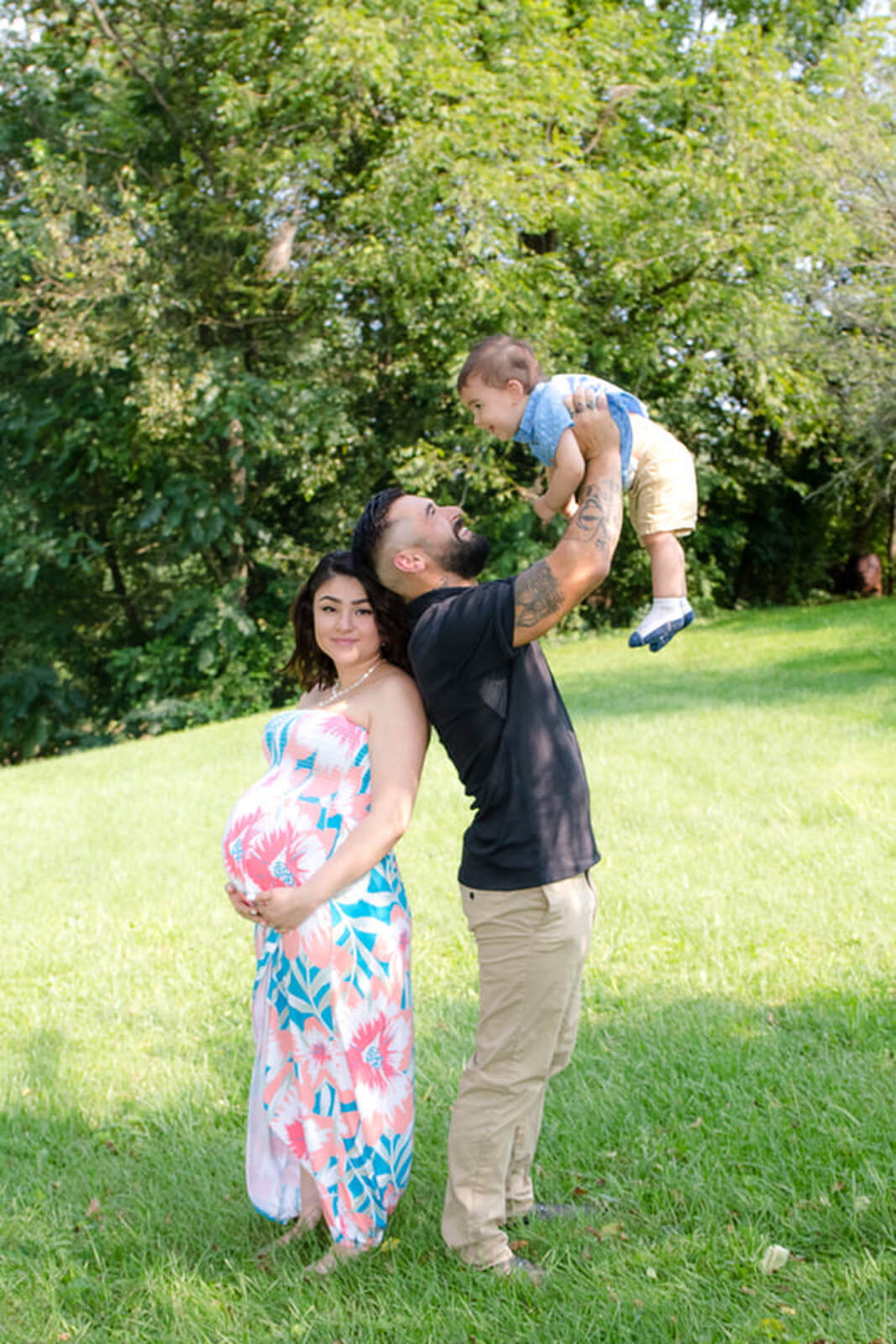 Family maternity, mom back to back to dad, dad holding up toddler, outdoors